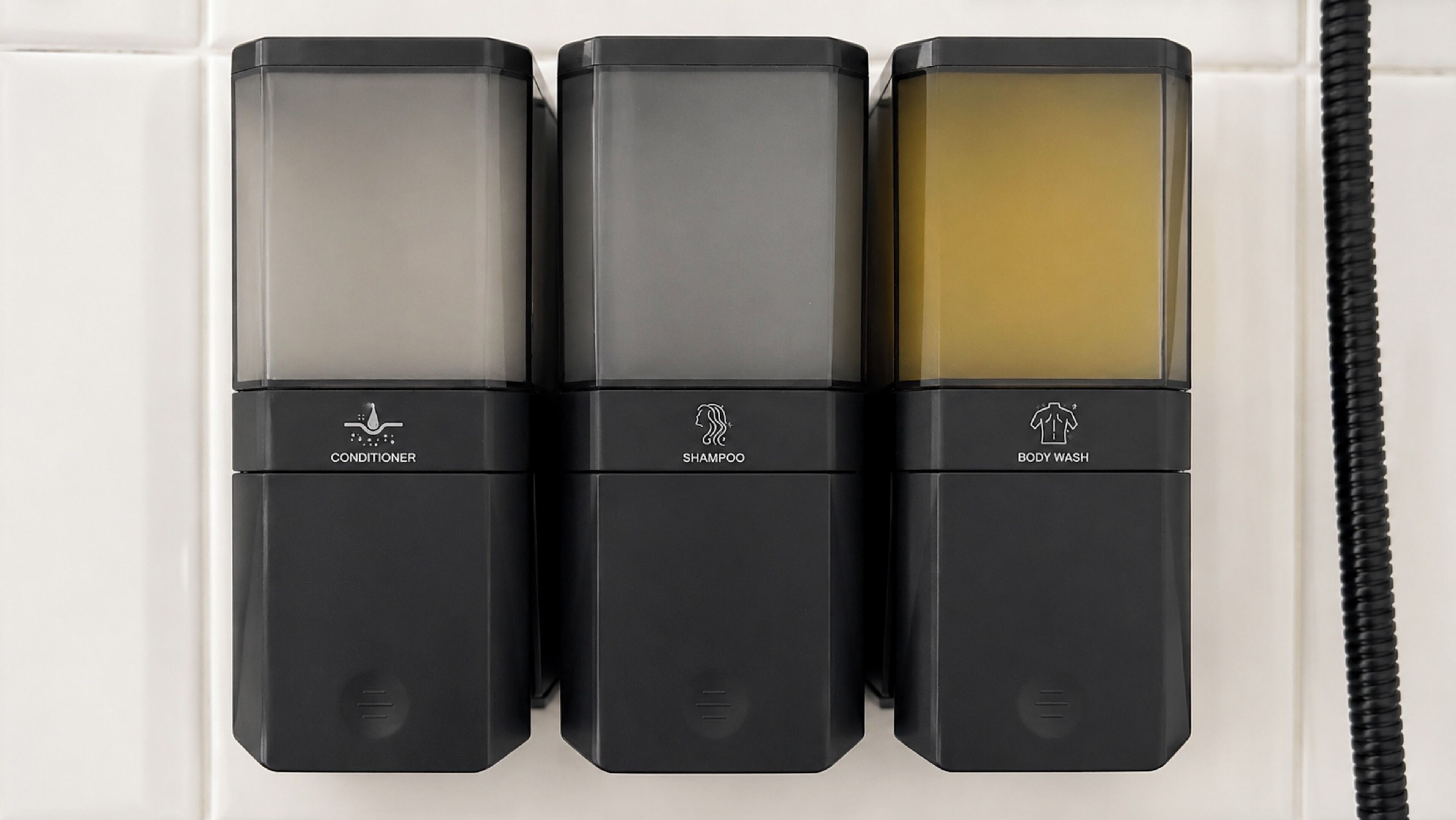 Three black dispensers labeled conditioner, shampoo, and body wash against a white tiled wall.