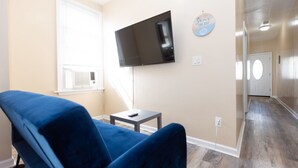 Ein Wohnzimmer mit einem blauen Sofa, einem flachbildfernseher an der Wand, einem kleinen Tisch und einem Flur, der zu einer Tür führt.