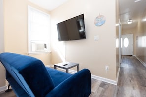 Ein Wohnzimmer mit einem blauen Sofa, einem flachbildfernseher an der Wand, einem kleinen Tisch und einem Flur, der zu einer Tür führt.
