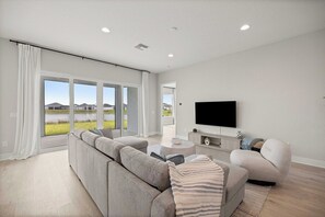 Ein modernes Wohnzimmer mit einem großen Ecksofa, einem Flachbildfernseher und einer Glastür, die einen Blick auf einen See und Berge freigibt.