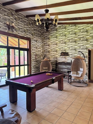 Une table de billard recouverte d’un feutre violet, une chaise en osier et un luminaire suspendu dans une pièce aux poutres apparentes et dont un mur est décoré d’un motif en briques.