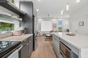 Eine moderne Küche mit Einbauofen, Kühlschrank und Kaffeemaschine. Ein Essbereich mit Tisch und Stühlen sowie ein Blick auf einen Garten durch ein Fenster.