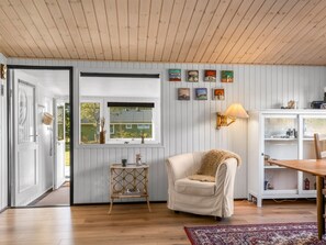 A room with a wooden floor, a white chair, a small table, a lamp, and a window with a view of a house. - "Into" - 490m from the sea by Interhome (Sæby)