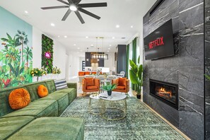Ein modernes Wohnzimmer mit einem grünen Sofa, orangefarbenen Sesseln, einem Kamin, einem Fernseher und einem Wandgemälde.