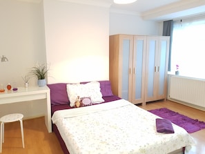 Ein Schlafzimmer mit einem Bett, einem Schreibtisch, einem Kleiderschrank, einem Fenster mit Vorhängen und einer Heizung.