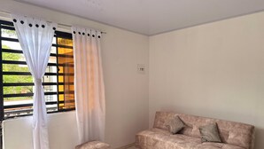 A room with a sofa, a small ottoman, a window with curtains, and a view of greenery outside. - Cozy country house with swimming pool and comfort only 5 min. from the city. (Villavicencio)