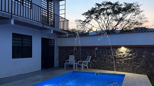 A swimming pool with water jets, a stone wall, and two white chairs. - Cozy country house with swimming pool and comfort only 5 min. from the city. (Villavicencio)