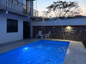 A swimming pool with water jets, a stone wall, and two white chairs.