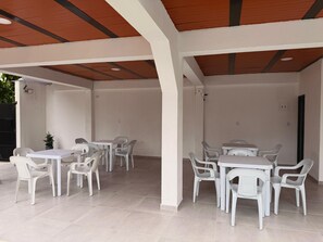 A covered outdoor seating area with white plastic tables and chairs, a tiled floor, and a wooden ceiling.