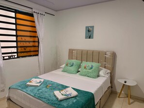 Una cama bien hecha con una colcha color turquesa y almohadones decorativos, una mesita de noche pequeña y una ventana con persianas.
