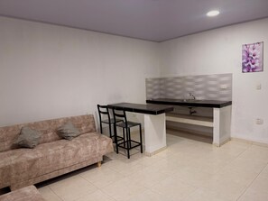 Una kitchenette con una encimera y un lavabo, un sofá con cojines, y una lámpara fijada en la pared.