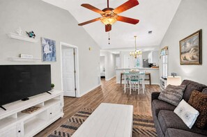 A modern living room with a flat-screen TV, a ceiling fan, and a dining area with a table and chairs. - Lowcountry Fairway Breeze at True Blue Golf n Pool (Pawleys Island)