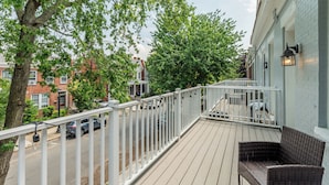 Ein Balkon mit weißer Reling, Korbmöbeln und Blick auf eine Straße mit parkenden Autos und Gebäuden.