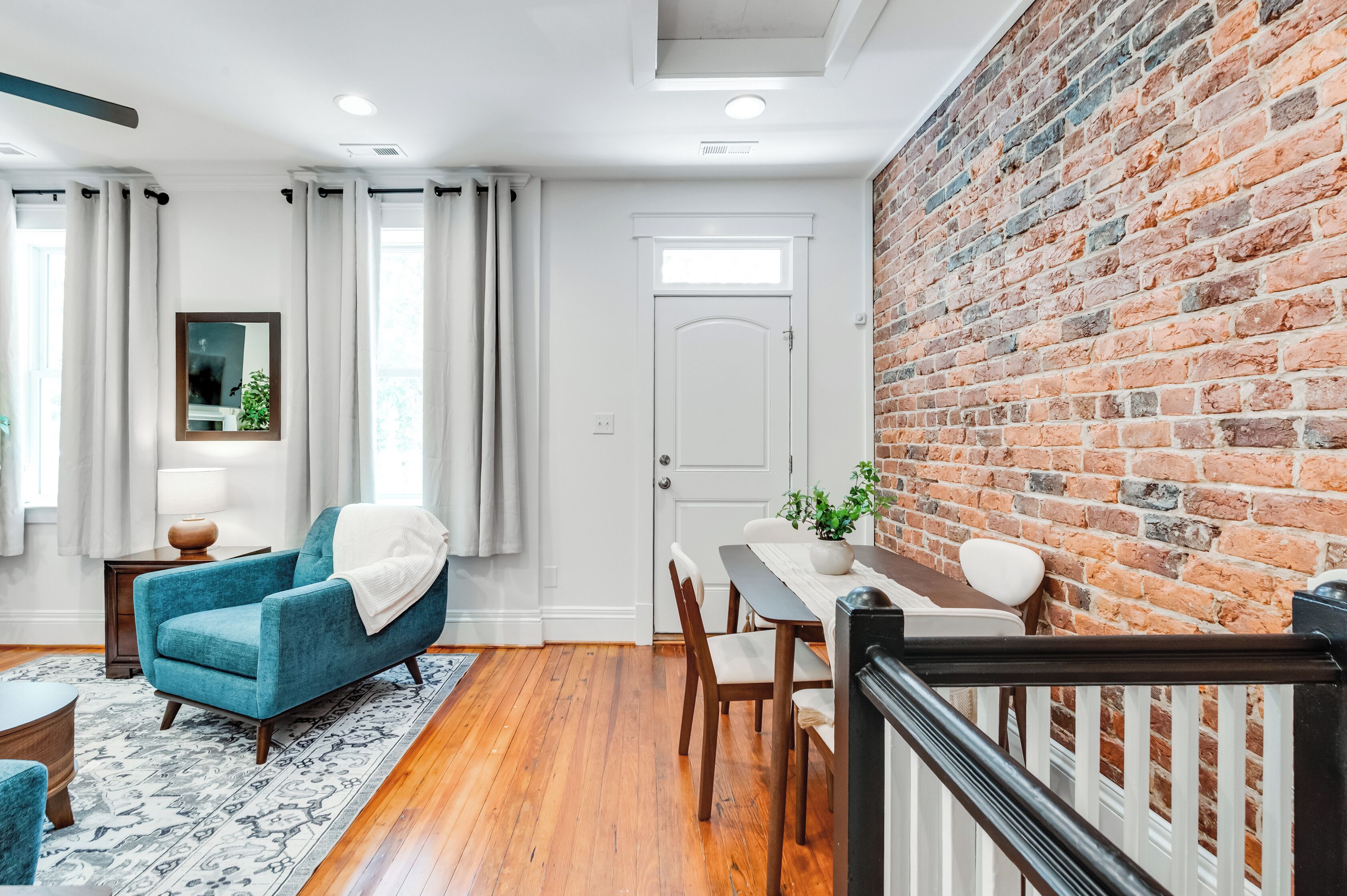 Une pièce avec un mur en briques, une table à manger avec des chaises, un fauteuil bleu, une porte blanche et une fenêtre avec des rideaux.