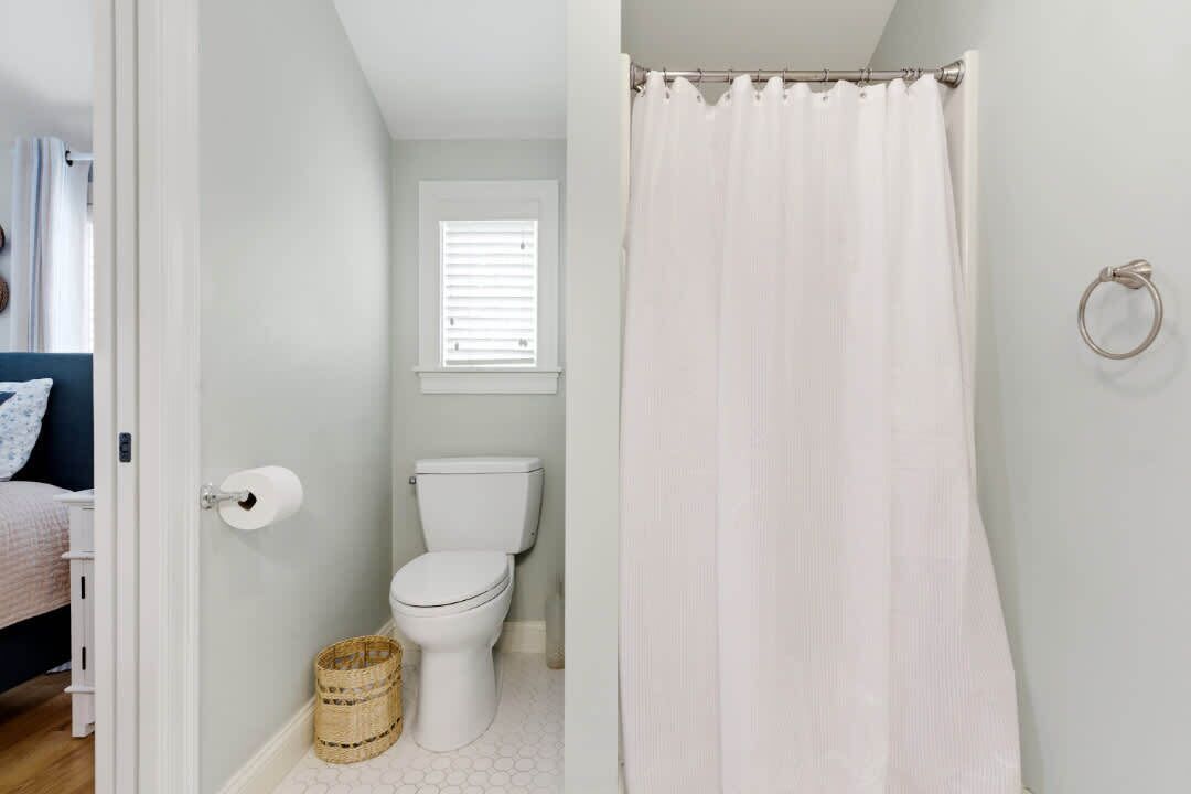 A bathroom with a toilet, a white shower curtain, a wicker basket, and a window with blinds.