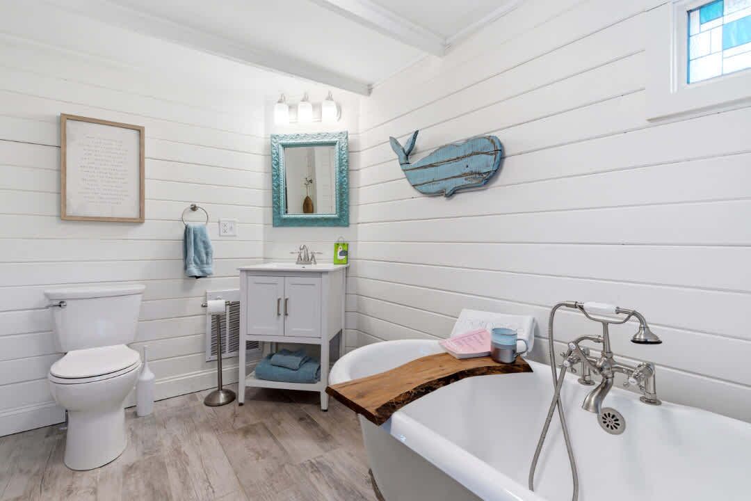 A bathroom with a white toilet, a sink with a mirror, a bathtub with a wooden tray, and a decorative whale on the wall.