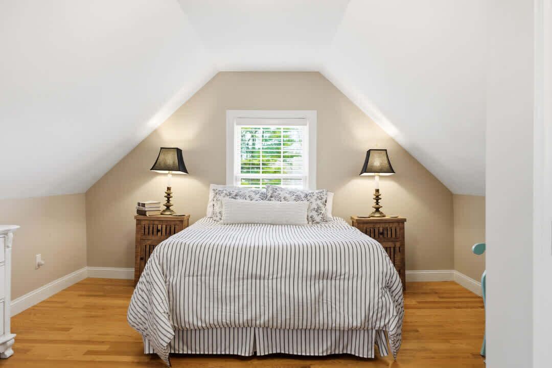 Un dormitorio con techo inclinado, una cama con ropa de cama a rayas, dos mesitas de noche con lámparas, una ventana con persianas y un aparador blanco.