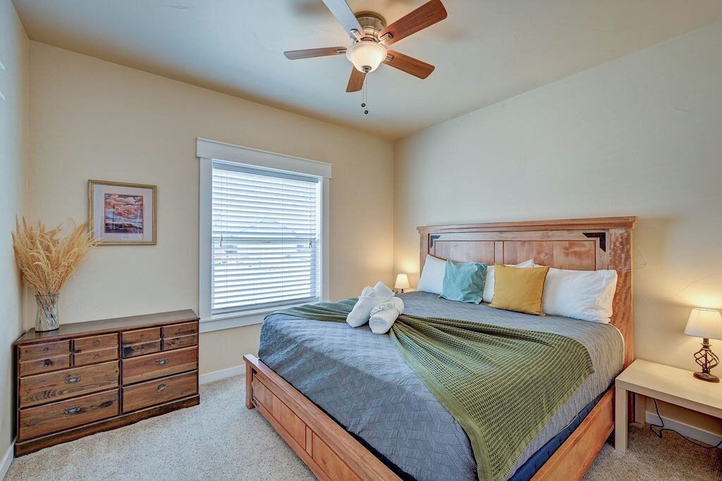 A bedroom with a bed, a ceiling fan, a window with blinds, a bedside table with a lamp, and a dresser.
