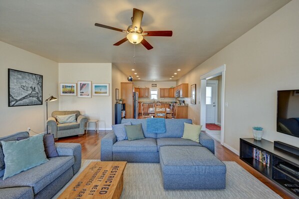 A living room with a sectional sofa, a ceiling fan, and a kitchen in the background.