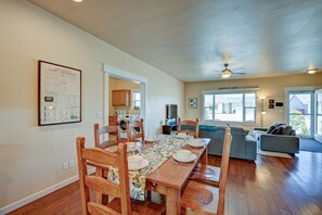 A dining area with a wooden table and chairs, a kitchen in the background, and a living area with a sofa and a ceiling fan.
