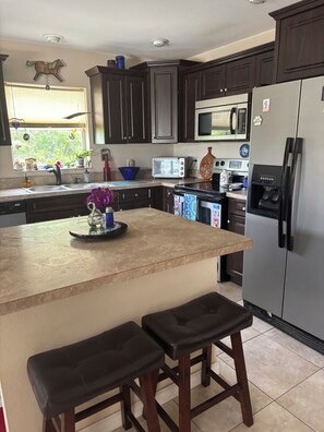 A kitchen with dark wooden cabinets, a stainless steel refrigerator, a microwave, and a toaster. There is a window with blinds and a plant on the windowsill.