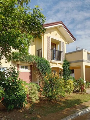 A two-story house with a balcony, surrounded by greenery and flowering plants.