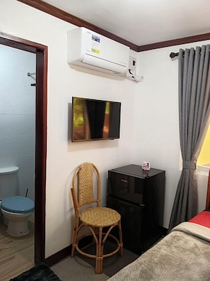 A room with a television mounted on the wall, a small black cabinet, a wicker chair, a toilet, and a bed with a grey curtain. - Noah Bed&Breakfast 3 PrivateRoom NEWHOME A Quiet Place to Stay Solar no Blackout (Puerto Princesa City)
