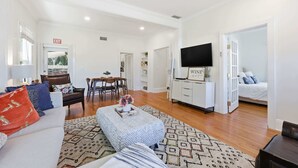A spacious living room with a sofa, ottoman, and coffee table, a dining area with chairs, a TV mounted on a white cabinet, and a bedroom with a bed and nightstand. - Porch on Pine Downtown (St. Augustine)