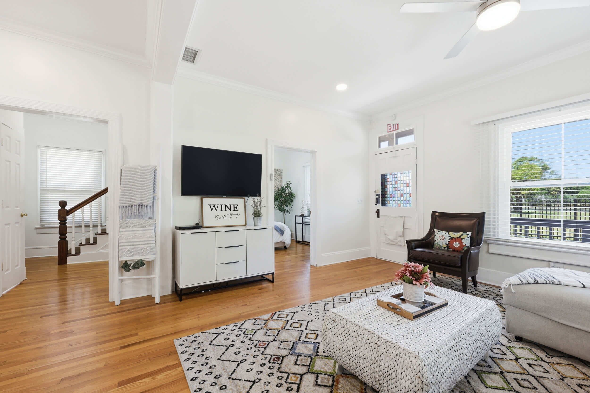 A spacious living room with a staircase, a flat-screen TV, a wine sign, a large window, and a view of the outside.