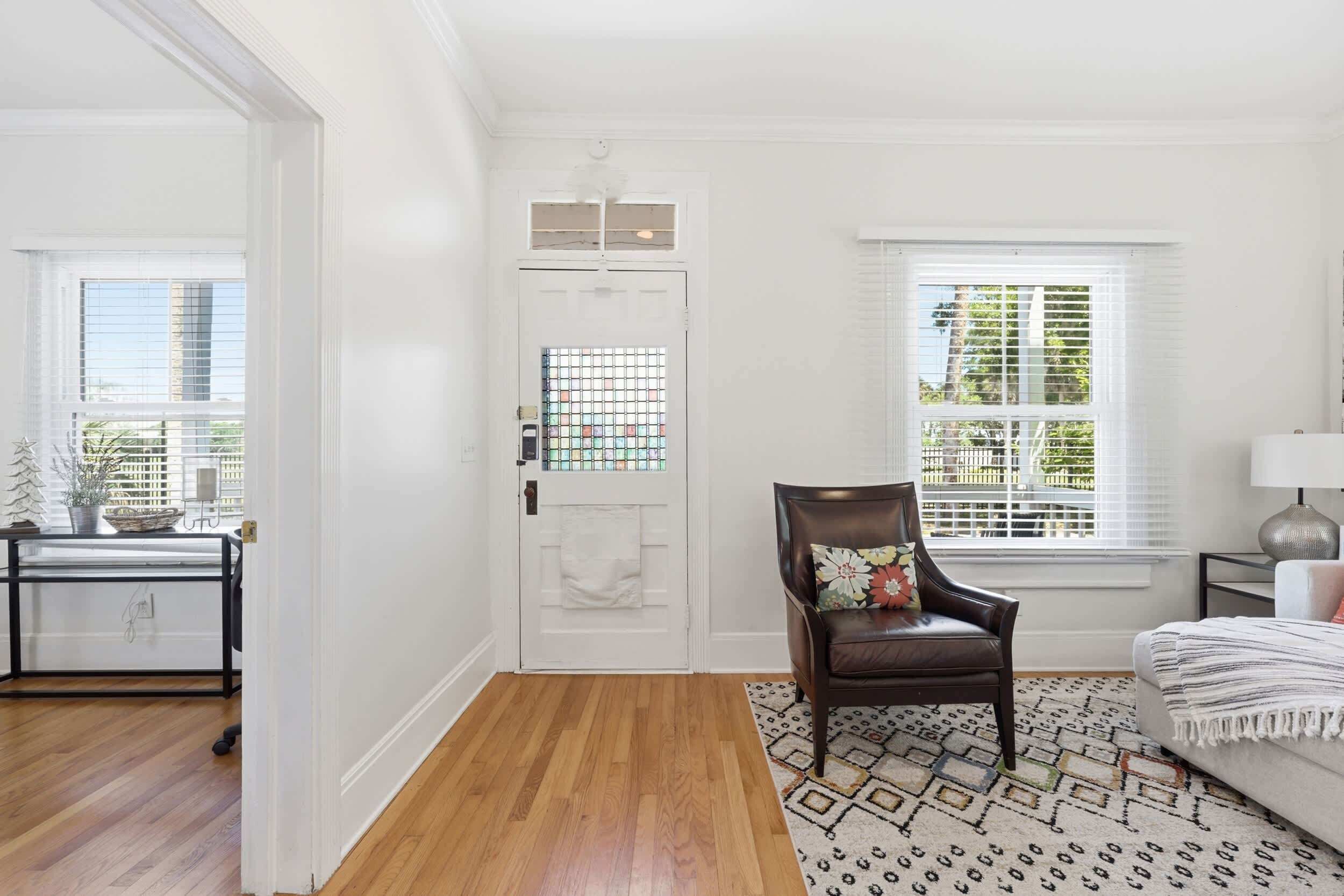 A room with a wooden floor, a white door with a window, a brown leather chair, a patterned rug, and a bed with a white headboard.