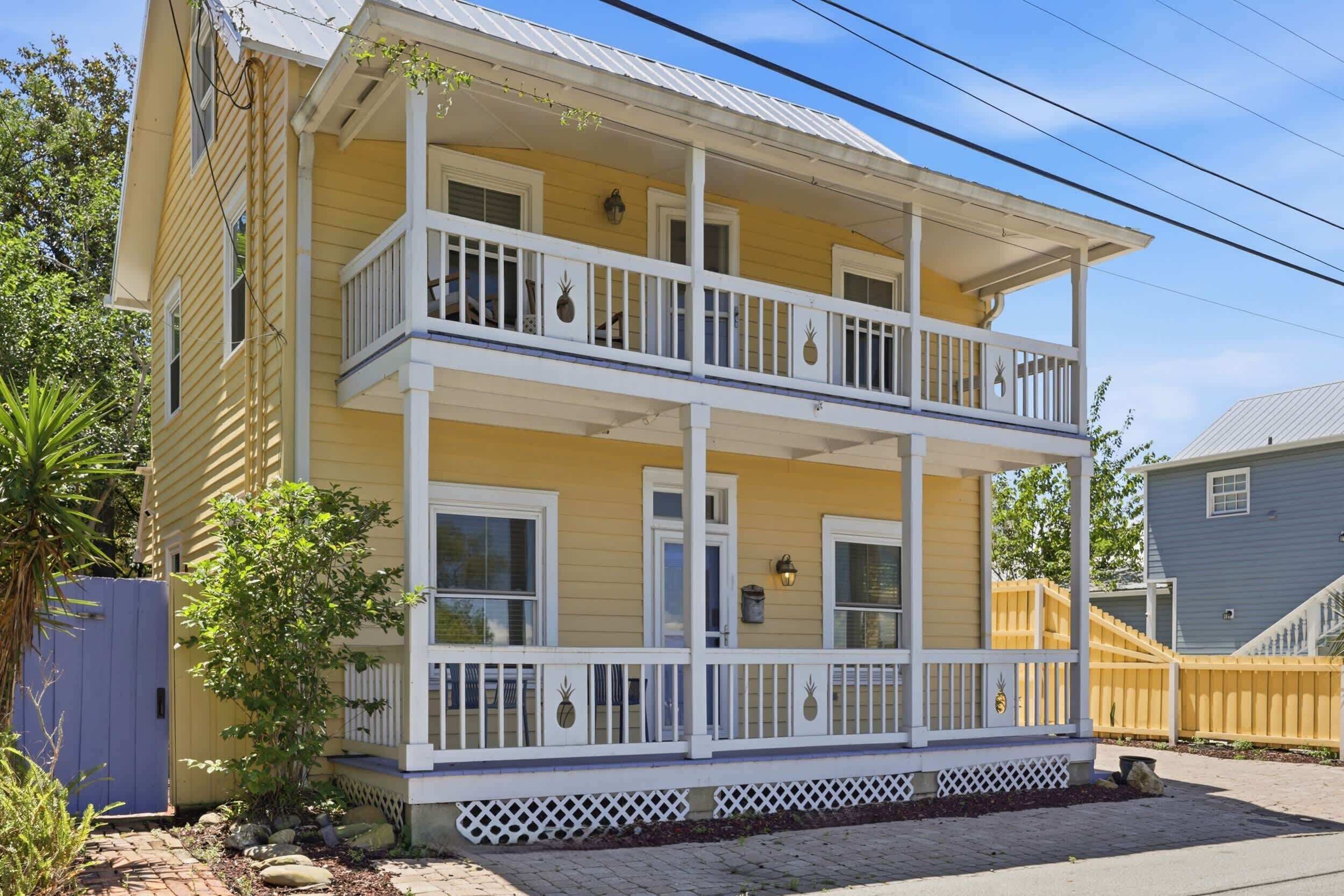 A two-story yellow house with a balcony, a wooden fence, and a small garden.