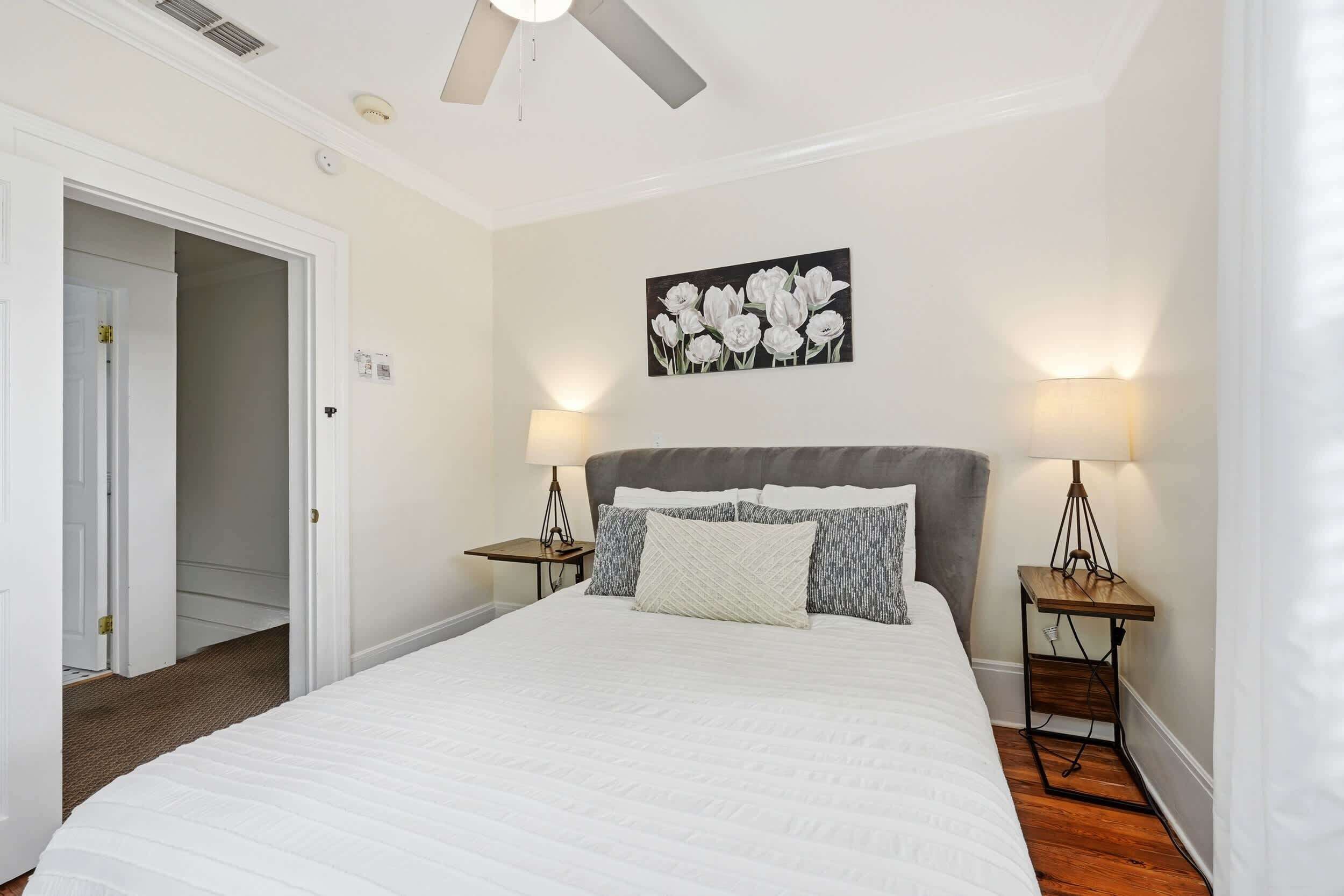 A bedroom with a large bed, two bedside tables with lamps, a ceiling fan, and a door leading to another room.