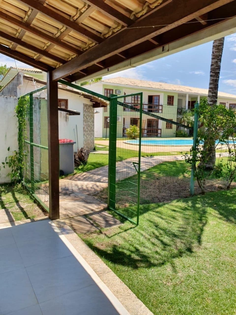 A covered outdoor area with a pool, a green fence, and a building in the background.
