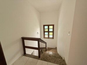 A narrow stairway with a wooden handrail, a small window with a view, and a light switch on the wall. - CASA 4 QUARTOS (Porto Seguro)