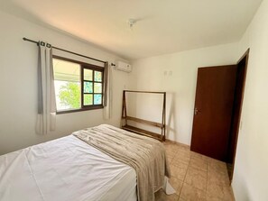 A bedroom with a bed, a window with curtains, an air conditioning unit, a wooden door, and a tiled floor. - CASA 4 QUARTOS (Porto Seguro)