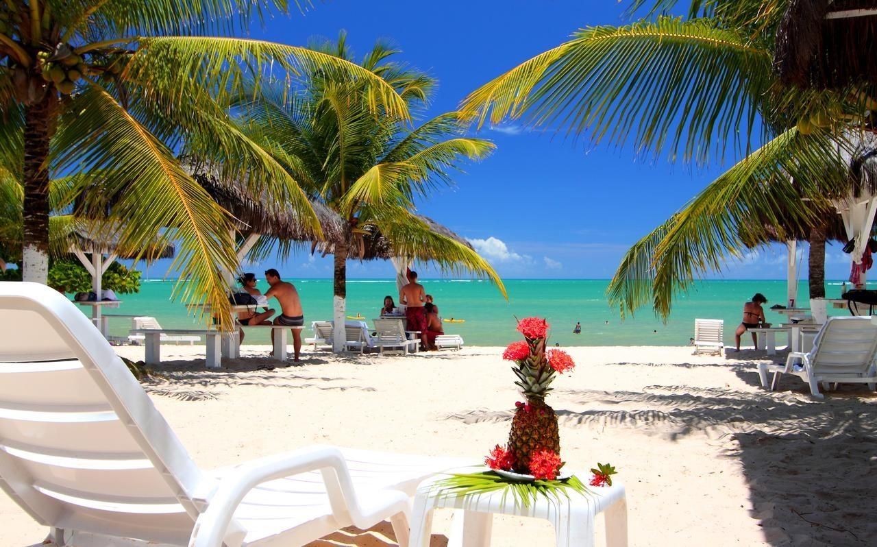 Une plage avec des chaises longues blanches, une table ornée d’un ananas et de fleurs, et des personnes à l’arrière-plan.