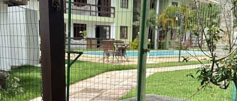 A view through a wire fence to a building with a green door, a concrete bench, and a grassy area with a pathway leading to a pool.