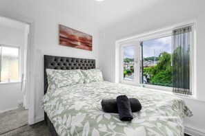 A bedroom with a bed, a window with a view, a painting on the wall, and a door. - Elegant 3 bedroom | 2 Bath | In Burnley (Lancashire)