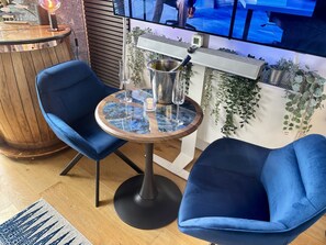 Ein modernes Zimmer mit einem runden Tisch, zwei blauen Stühlen, einer Holzwand als Blickfang und einem Fernseher, der über dem Tisch angebracht ist.