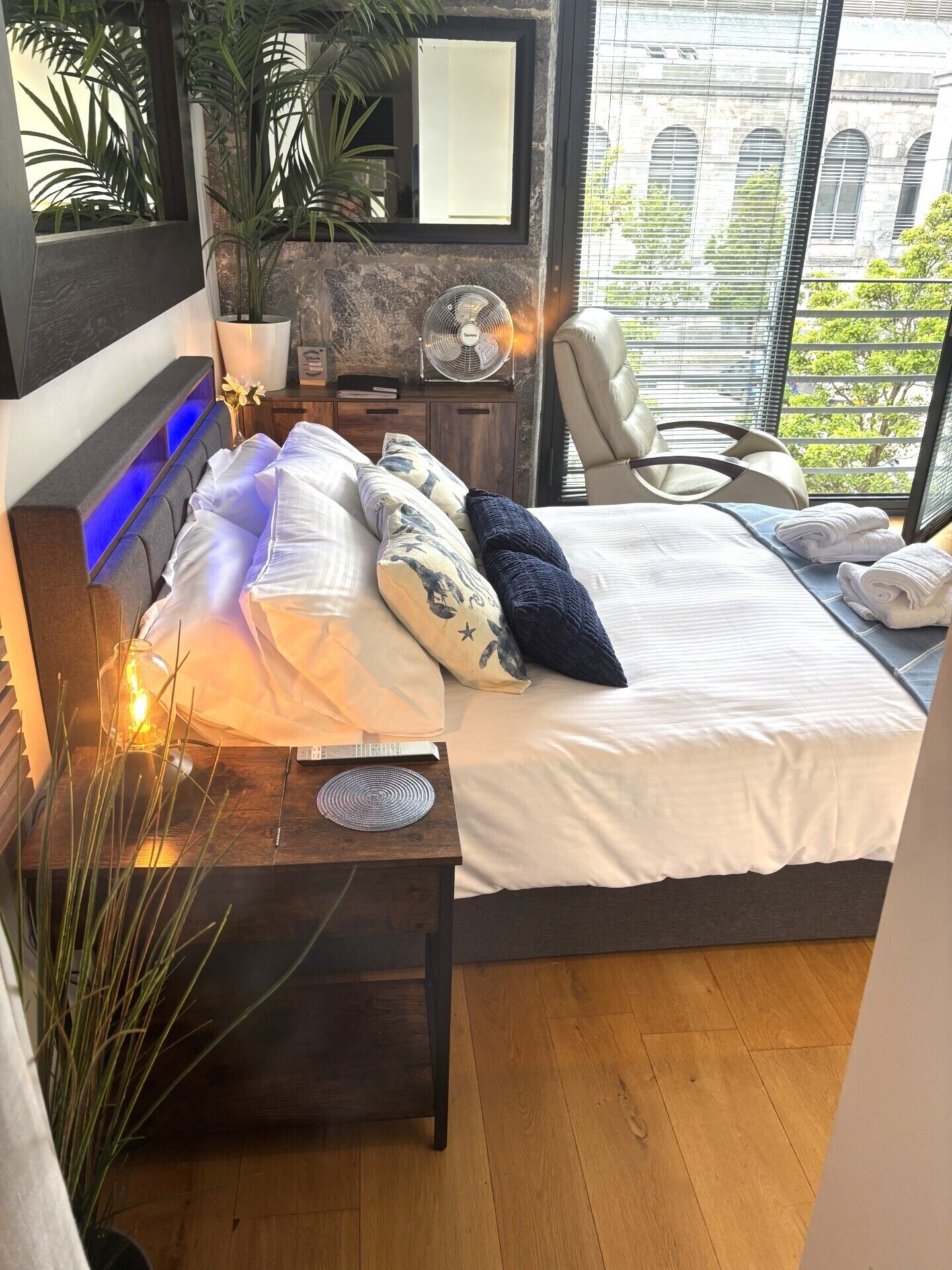A modern bedroom with a bed, bedside table, lamp, and a view of a building through large windows.