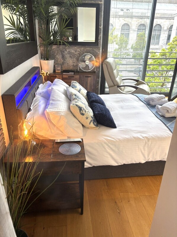 A modern bedroom with a bed, bedside table, lamp, and a view of a building through large windows. - Luxury studio accommodation, located in historic Royal William Yard, Plymouth. (Plymouth)