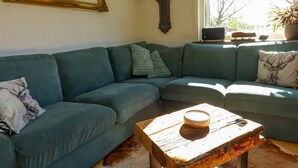 Uma sala de estar com sofá seccional, mesa de centro de madeira, relógio na parede e janela com vista.