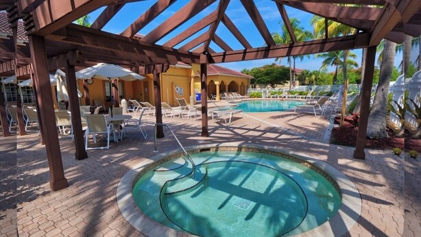 A pool area with lounge chairs and umbrellas, a covered seating area, and a pool with a circular design. - Peaceful Beachwalk Escape! Updated 3rd-Floor Condo w/ Lanai, Pool & Hot Tub (Fort Myers)