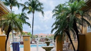 A courtyard with a central fountain, palm trees, and a pool in the background. - Peaceful Beachwalk Escape! Updated 3rd-Floor Condo w/ Lanai, Pool & Hot Tub (Fort Myers)