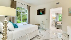 A bedroom with a bed, a TV, a window with curtains, a small table with a vase, and a hallway leading to another room.
