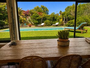 A wooden table with chairs, a potted plant, and a view of a swimming pool and garden.