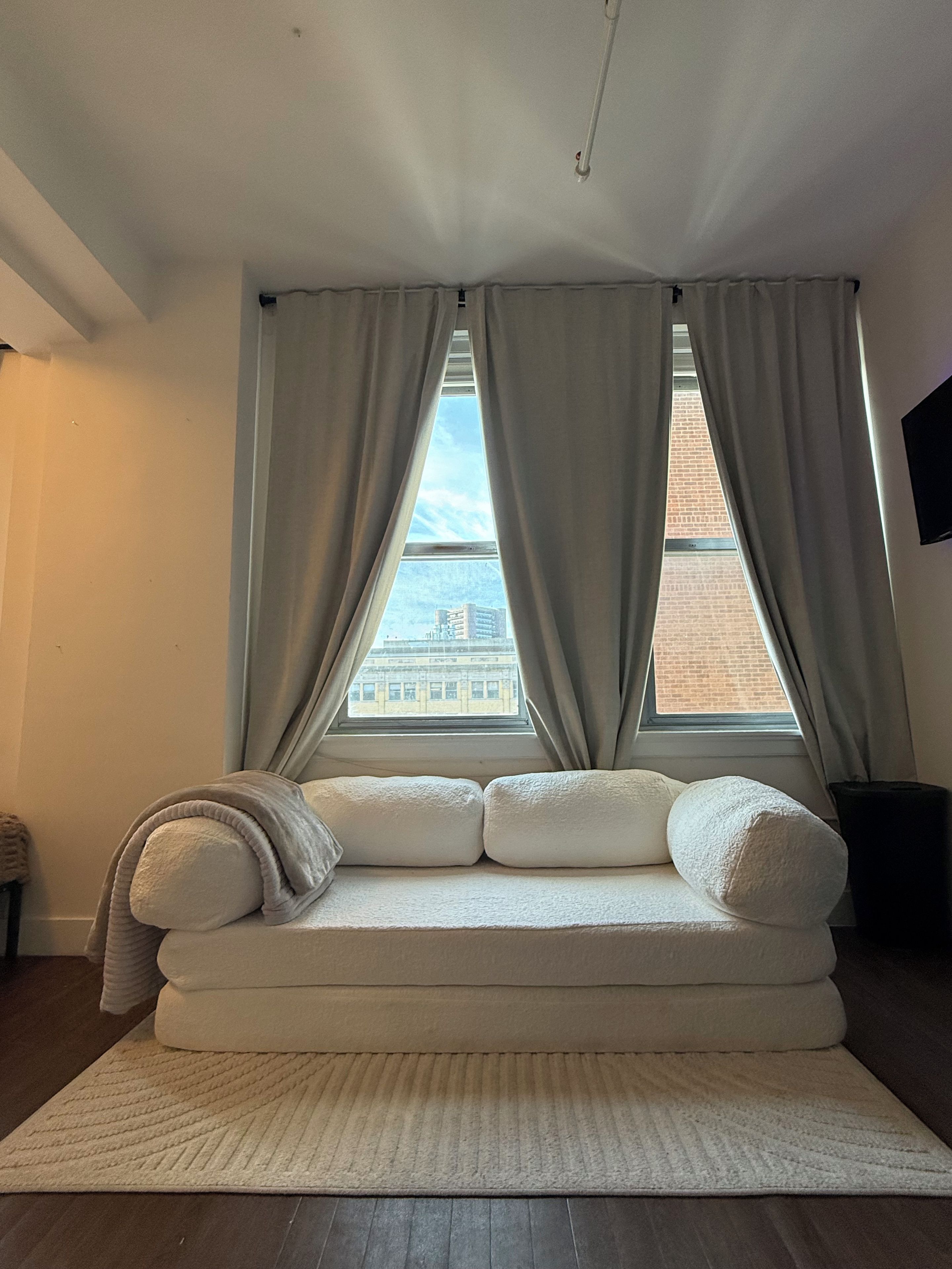 A room with a large window, a white sofa with cushions, a rug, and a television on the wall.
