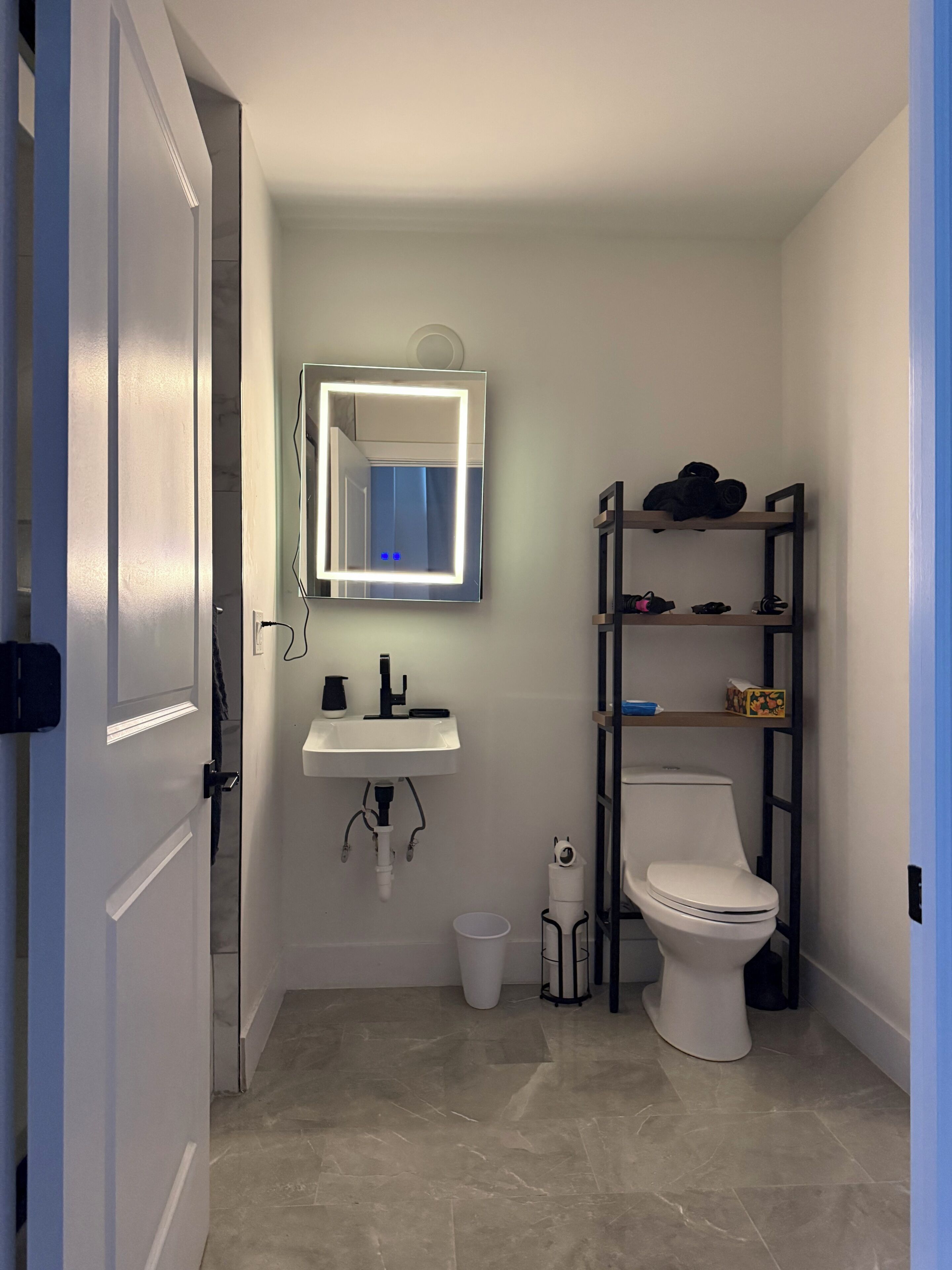 A bathroom with a white sink, a toilet, a mirror with lights, and a shelf with items.