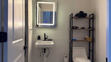 A bathroom with a white sink, a toilet, a mirror with lights, and a shelf with items.