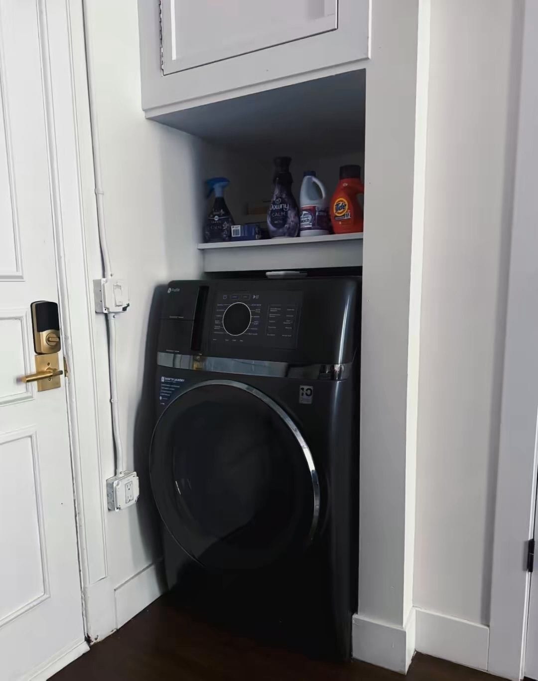 Un lavadero pequeño con lavadora de carga frontal, un estante con diversos productos de limpieza y una puerta con cerradura.